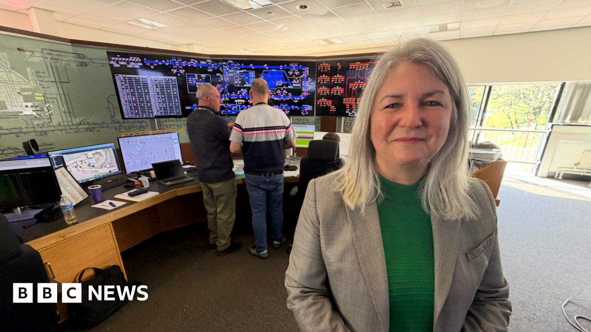 Sarah McManus, who is wearing a green jumper and a grey blazer, stands in front of the soon to be decommissioned system at the control room in South Gosforth. In the background are two controllers who are discussing how the network is currently operating.