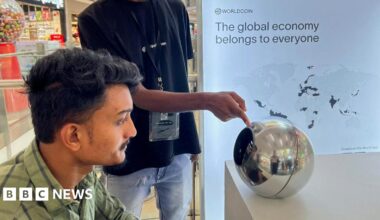 A man in India sitting before an earlier World device, a silver orb, to have his eye scanned as a form of human identification. He has dark hair and a green shirt, and looks straight into the orb, on a desk in front of him, while another man points to it.