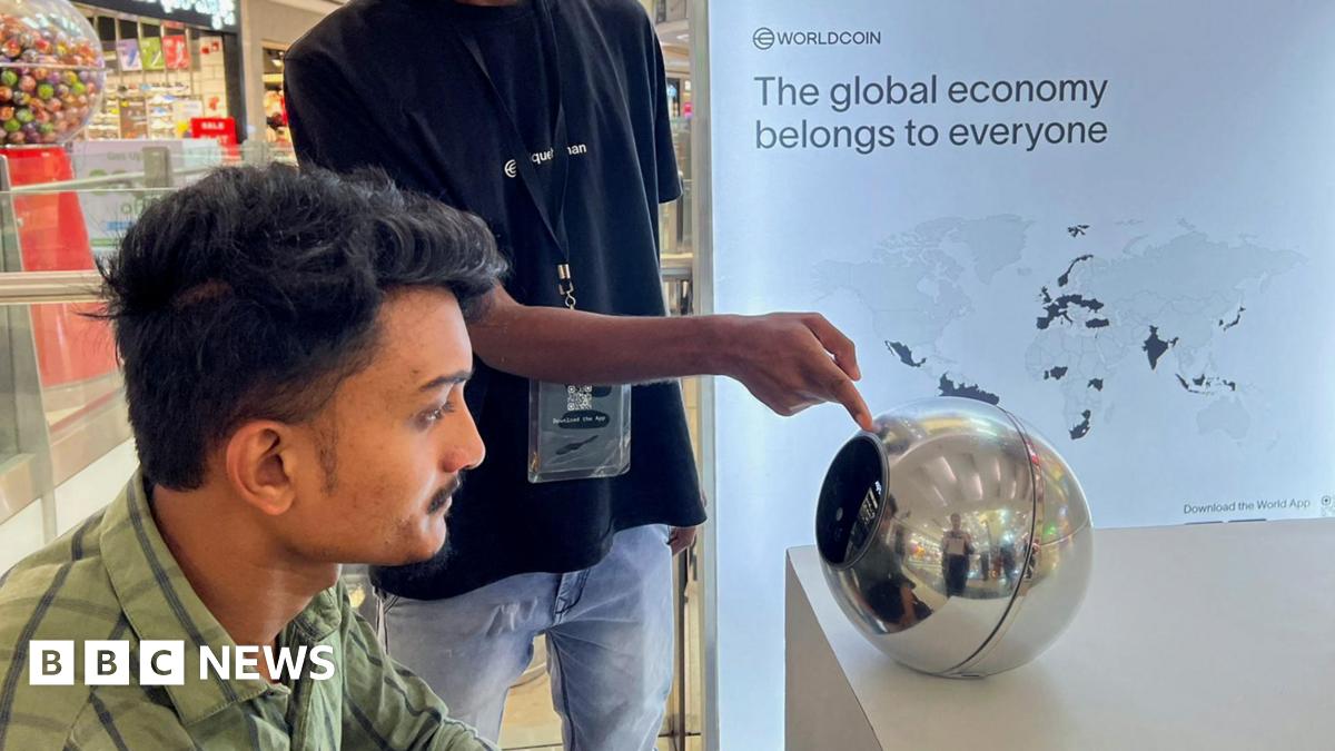 A man in India sitting before an earlier World device, a silver orb, to have his eye scanned as a form of human identification. He has dark hair and a green shirt, and looks straight into the orb, on a desk in front of him, while another man points to it.