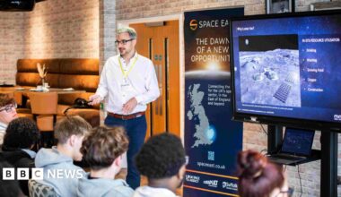 Andrew McCulloch, from Minima, giving a space engineering workshop to students at Suffolk New College. He is wearing a white shirt and navy trousers and addressing a group of students who are sitting down in front of him and a television screen.