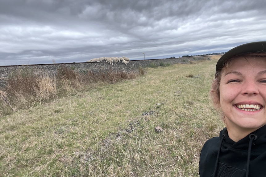 Young women smiles to camera. She is wearing a cap. There are a flock of sheep in the background.