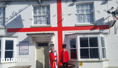 Two people dressed as an English knight and a Beefeater stood outside a white pub with a red stripe which makes it look like a English flag.