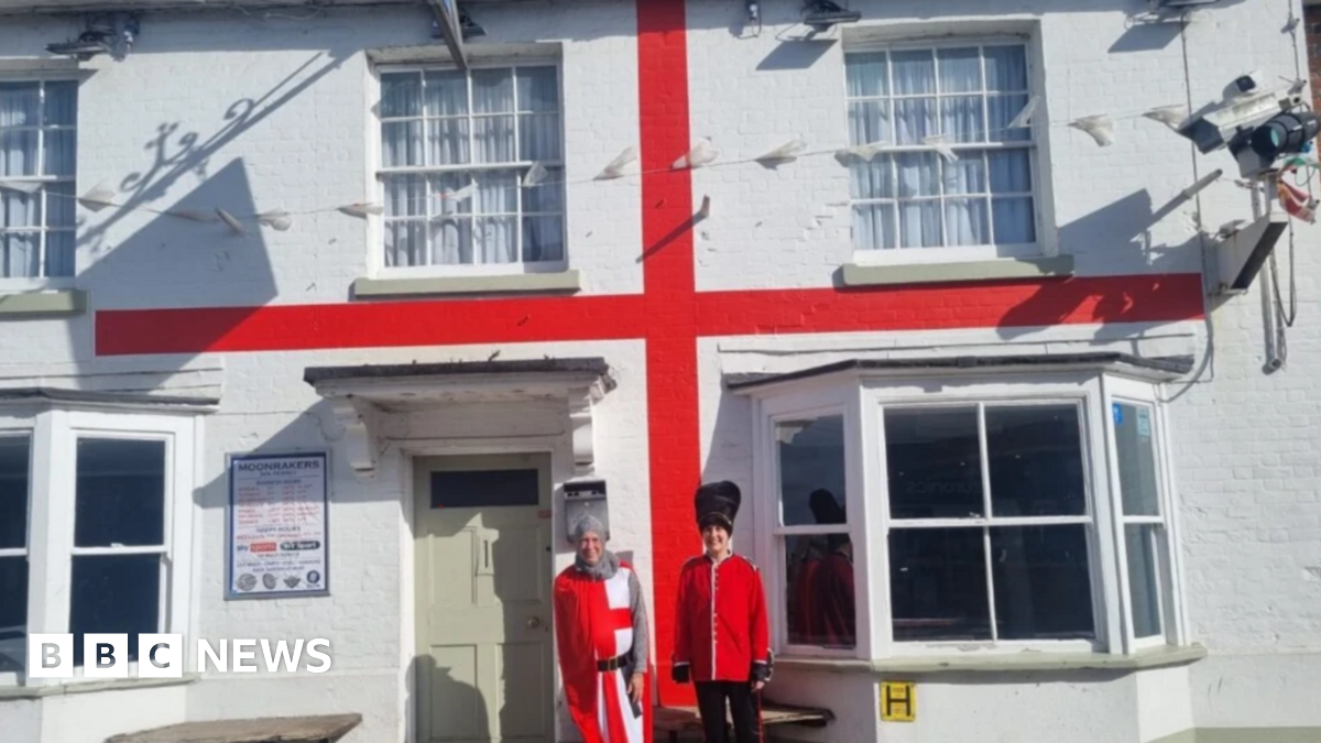 Two people dressed as an English knight and a Beefeater stood outside a white pub with a red stripe which makes it look like a English flag.