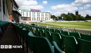 photograph taken from the stands of New Road showing green plastic seats in rows in front of the camera. A white building is pictured on the edge of the stadium with trees and Worcester Cathedral overlooking the pitch.