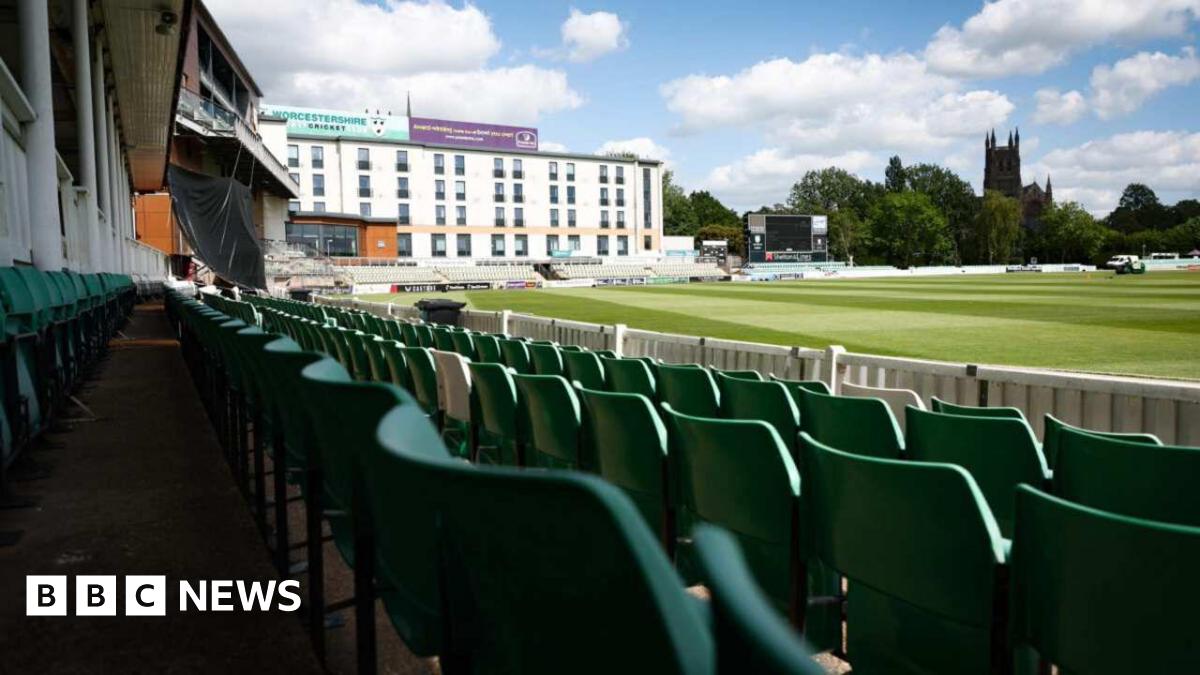 photograph taken from the stands of New Road showing green plastic seats in rows in front of the camera. A white building is pictured on the edge of the stadium with trees and Worcester Cathedral overlooking the pitch.