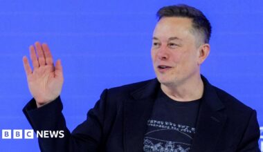 Elon Musk waving to a crowd from a stage wearing a black blazer and a black t-shirt.