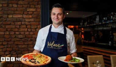 Jason Maimone smiles as he holds a pizza and another Italian dish in his restuarant. He wears a blue apron with the Italian tricolour over his chef whites.