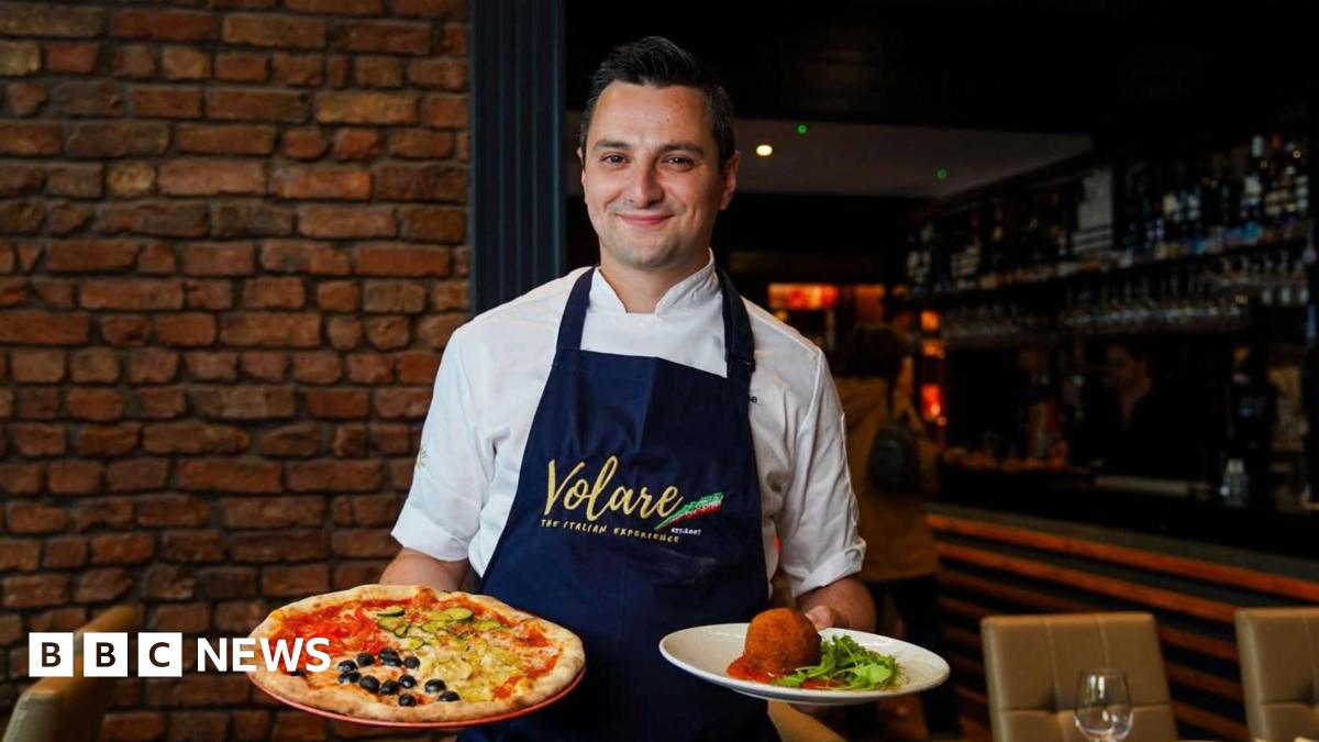 Jason Maimone smiles as he holds a pizza and another Italian dish in his restuarant. He wears a blue apron with the Italian tricolour over his chef whites.