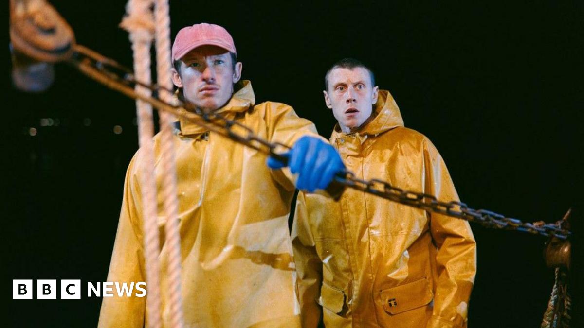 The image shows two men are dressed in bright yellow oilskin jackets. It is pitch black behind them. The man in the foreground is holding a chain and ship's ropes hang vertically in front of him. The second man has his mouth open in shock.
