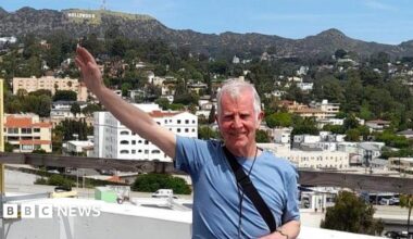 Jim, a mature man with white short hair and wearing a light blue T-shirt raises his arm towards the "Hollywood" sign as he checks out the roof on his tour of the Capitol Records building.