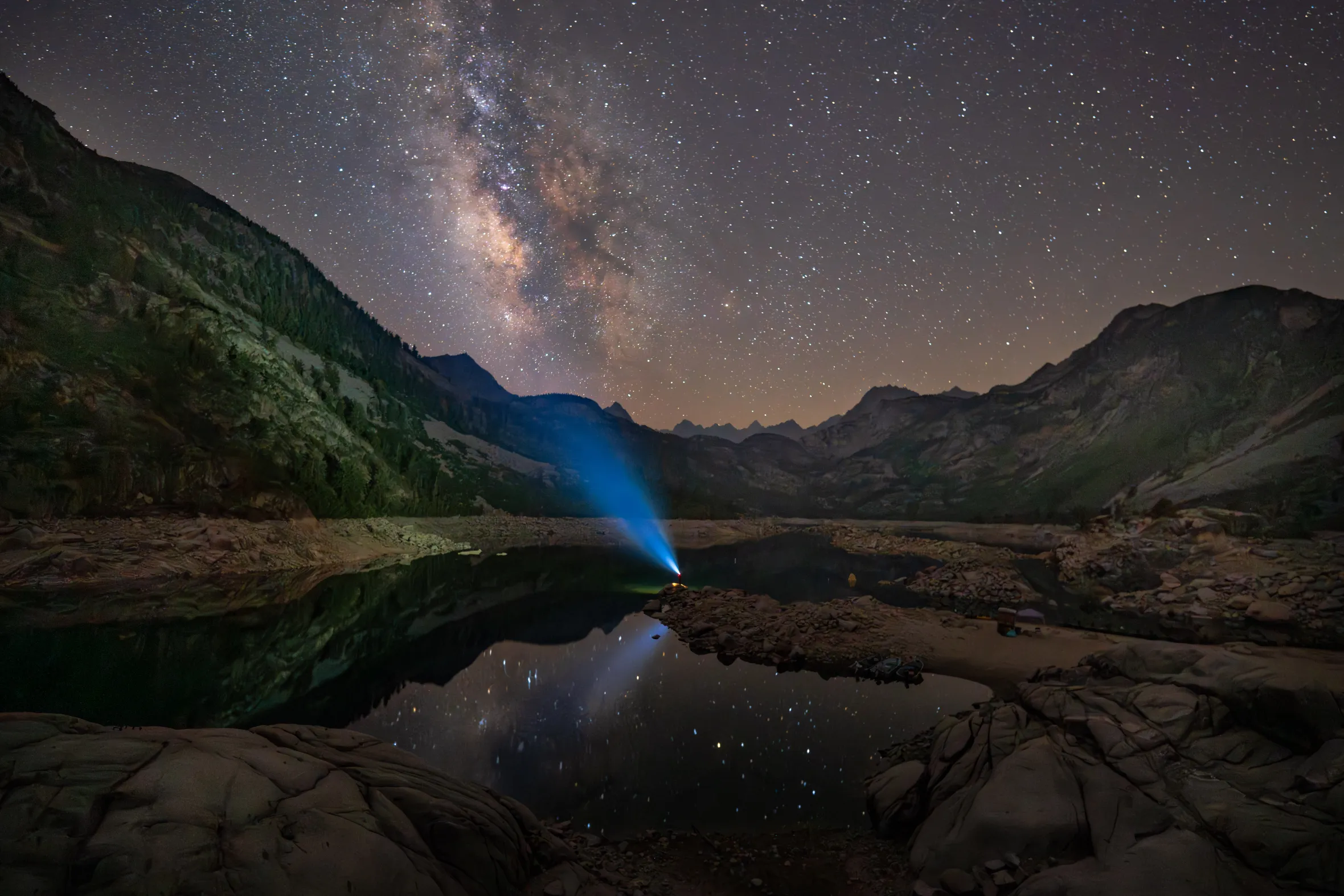A lone traveler's flashlight beam pierces the darkness at the edge of the glass-still Lake Sabrina in California’s Eastern Sierra, with the Milky Way visible above.