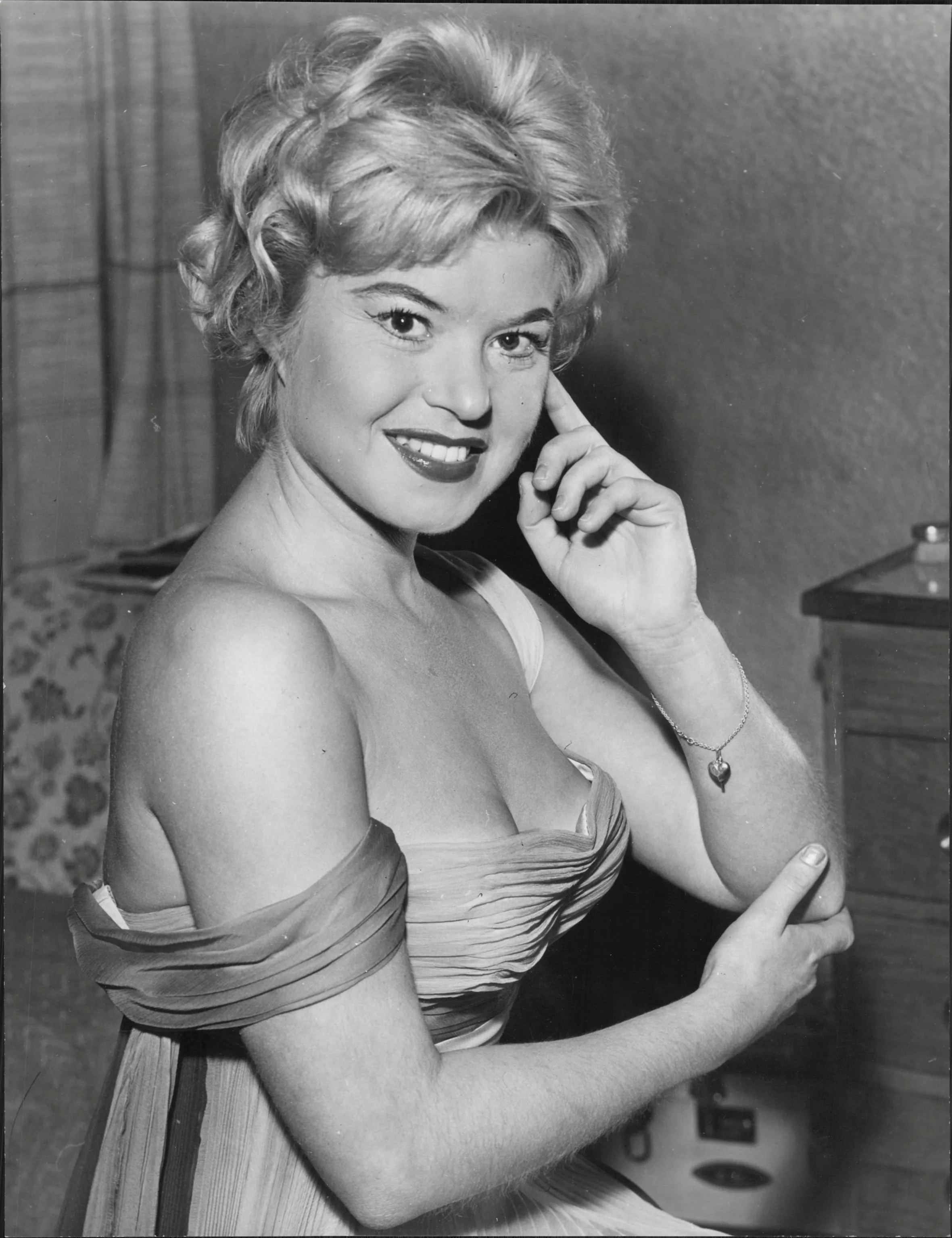 Shani Wallis, a smiling actress, with blonde hair, wearing an off-the-shoulder pleated dress.