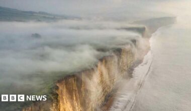 Thick fog rolls off the top of cliffs and into the sea. The sky is grey and foggy. The cliffs have a beige colour to them. A calm sea is rolling onto the shore at the foot of the cliffs.