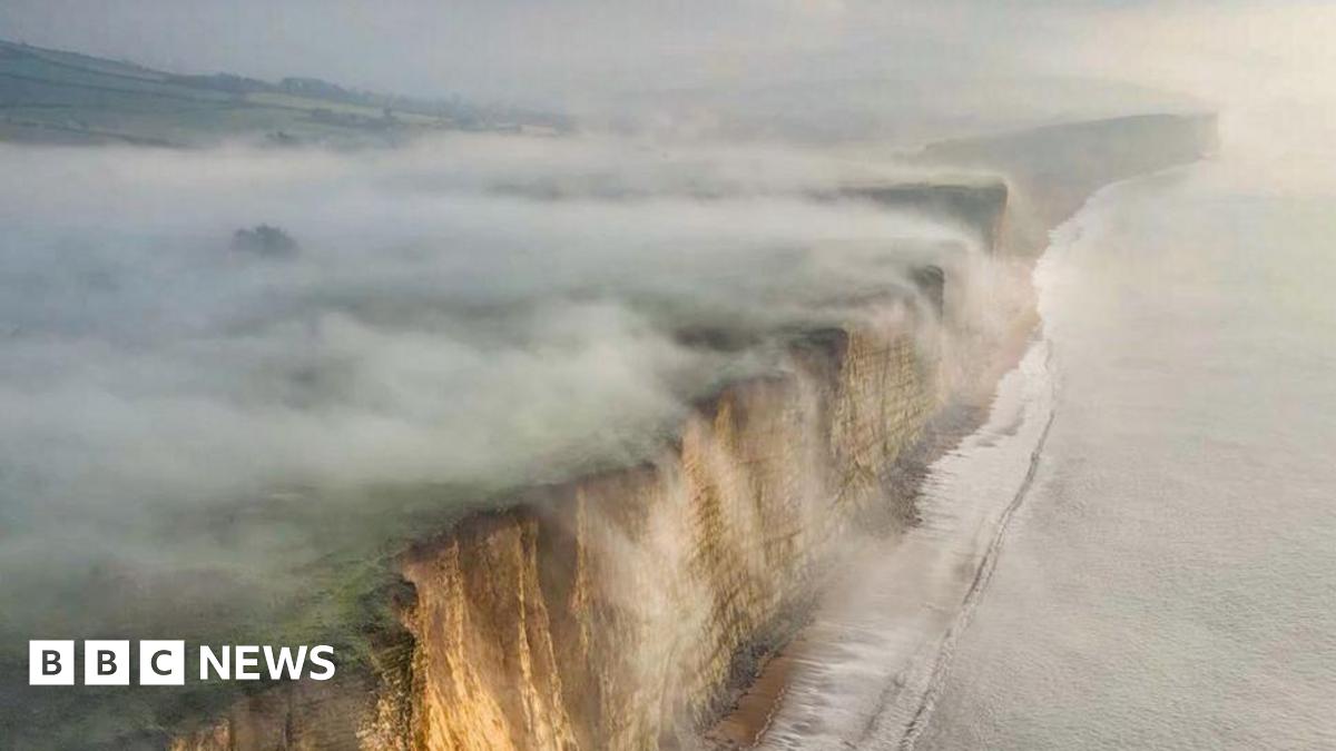 Thick fog rolls off the top of cliffs and into the sea. The sky is grey and foggy. The cliffs have a beige colour to them. A calm sea is rolling onto the shore at the foot of the cliffs.