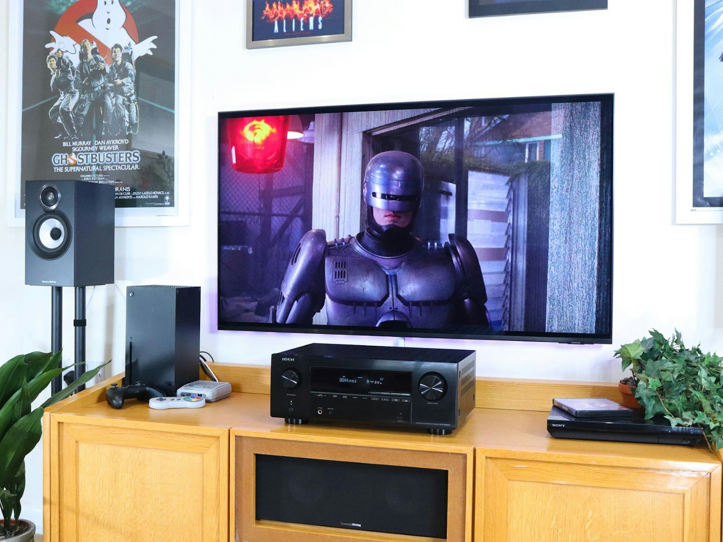 Denon AVR-X2800H Home Cinema Amplifier in a living room on a stand with Xbox and blu-ray player and speakers with a TV showing robocop