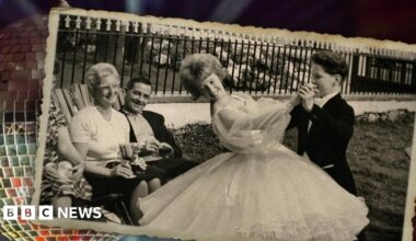 Black and white image from 1963 taken in the Isle of Man of junior dance champions June Batty in a white ballroom dress and John Halliday in a black tuxedo resting on a silver glitterball. They are doing an old-time tango pose while parents sitting on deck chairs look on smiling and Eva holding their winning trophies.