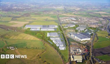 A computer generated image of the proposals shows large grey industrial units. A road separates the site from abother industrial sire and residential houses.