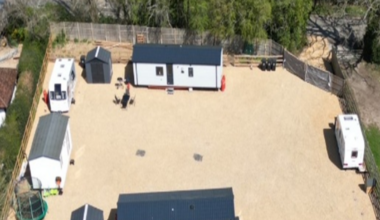 A view of the field. About six caravans are parked on hardstanding along with other vehicles and yellow diggers. There is a pylon in the middle of the field.