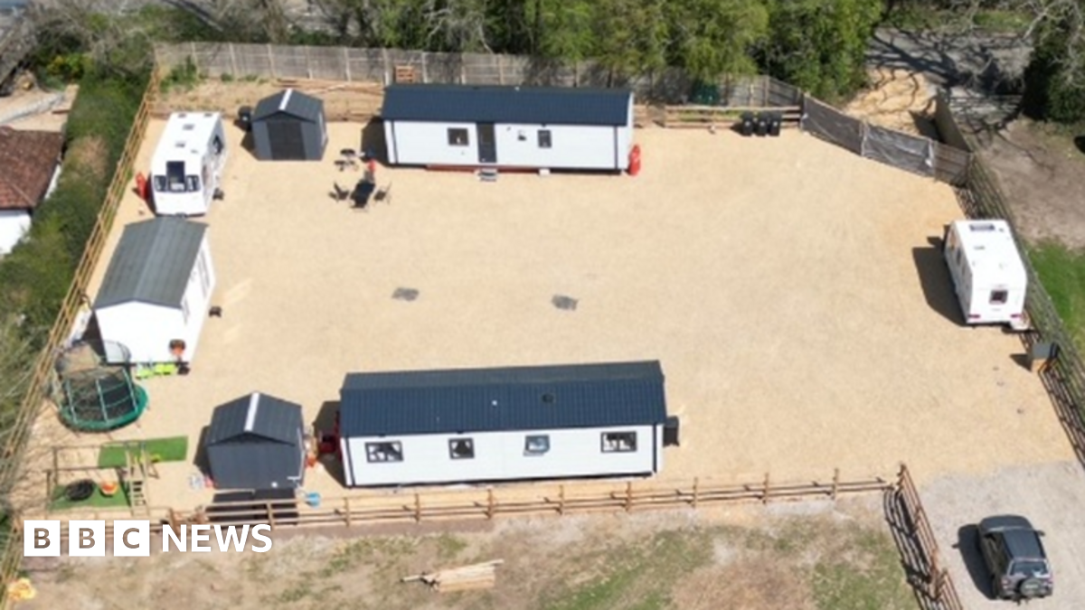 A view of the field. About six caravans are parked on hardstanding along with other vehicles and yellow diggers. There is a pylon in the middle of the field.