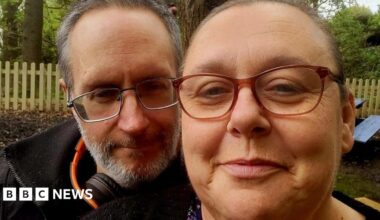 A man with grey hair and beard looks into the camera in a black jacket, orange and black headphones around his neck. He is beside a woman who also looks into the camera, hair tied back. Both wear glasses and have slight smiles on their faces. Behind them woodland, a fence and trees are visible.