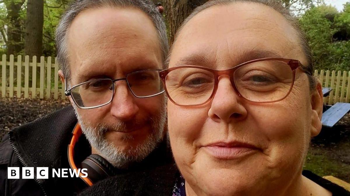 A man with grey hair and beard looks into the camera in a black jacket, orange and black headphones around his neck. He is beside a woman who also looks into the camera, hair tied back. Both wear glasses and have slight smiles on their faces. Behind them woodland, a fence and trees are visible.