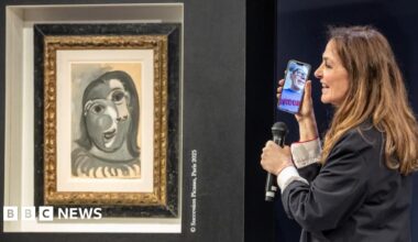 A blue painting by Picasso, in his trademark cubist style, framed on the wall beside a woman holding a microphone up to her mobile phone, which shows a video call with a man wearing glasses - both are smiling broadly.