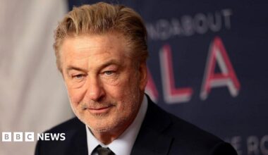 Alec Baldwin looks ahead in a suit and tie. His greying hair is slicked back and he is wearing a soft smile. In the background is a red carpet backdrop in which the word "gala" is visible.