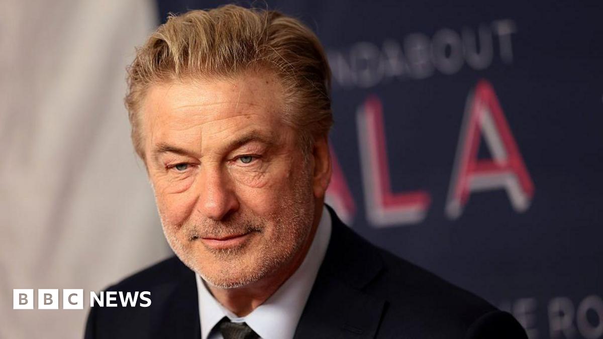 Alec Baldwin looks ahead in a suit and tie. His greying hair is slicked back and he is wearing a soft smile. In the background is a red carpet backdrop in which the word "gala" is visible.