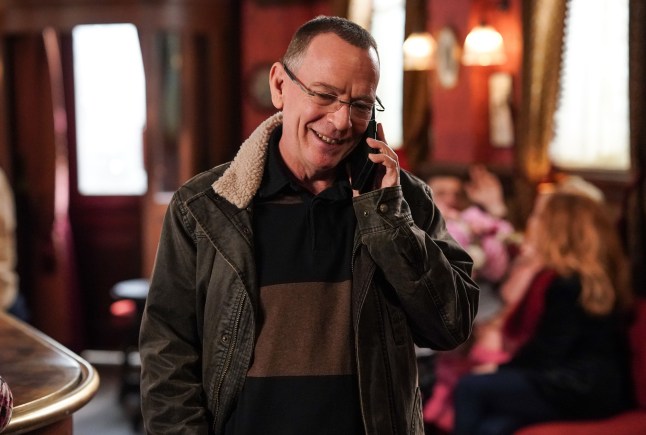 Ian Beale smiling while standing in the pub on his phone in EastEnders.