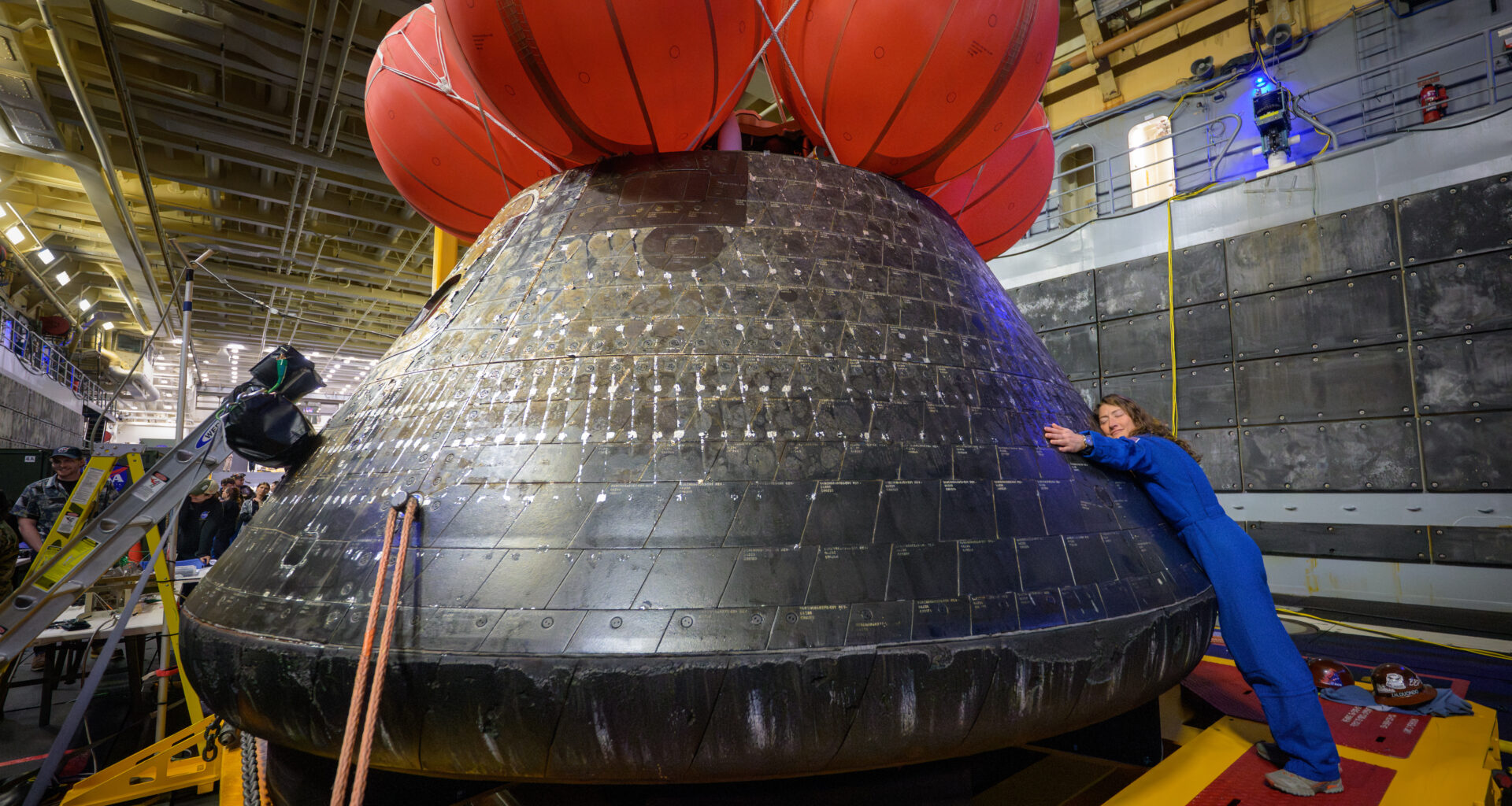 NASA astronaut Christina Koch hugs the Orion spacecraft with her face toward the camera. She is wearing a bright blue jumpsuit. Orion is a rounded cone shaped spacecraft. Its exterior is a sooty black. There are several large orange balloons attached to its top. Orion looks about twice Koch's height.