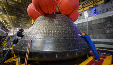 NASA astronaut Christina Koch hugs the Orion spacecraft with her face toward the camera. She is wearing a bright blue jumpsuit. Orion is a rounded cone shaped spacecraft. Its exterior is a sooty black. There are several large orange balloons attached to its top. Orion looks about twice Koch's height.