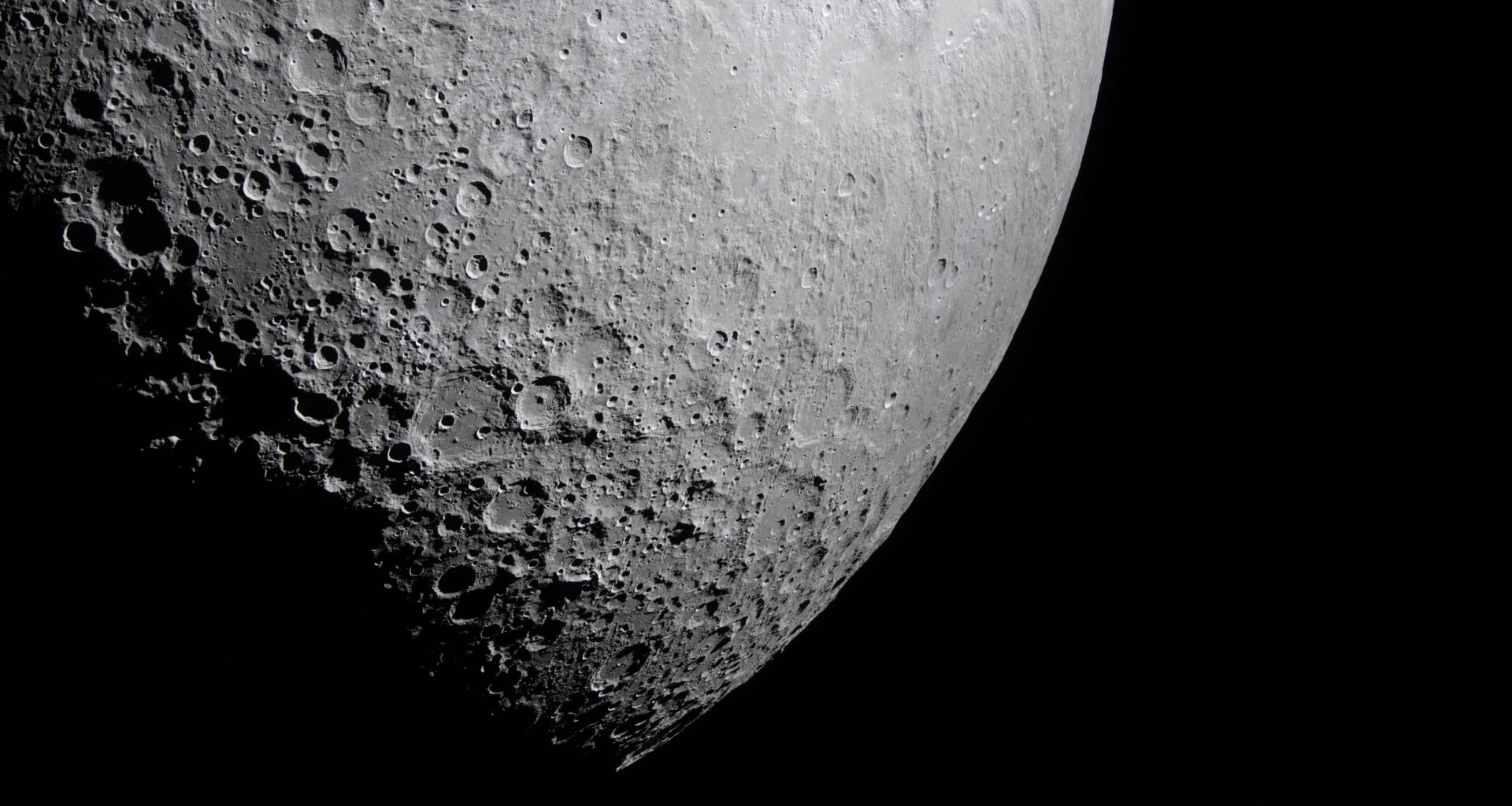 Part of the Moon stands out against the blackness of space. Its surface is marked with many craters of different sizes. The craters are mostly on the left and bottom. The upper right portion of the Moon's surface is much smoother.