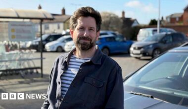 Gareth Wild stands in front of his car at the car park of his local supermarket. He looks at the camera and has dark brown hair and a beard. He is wearing a striped t-shirt underneath a navy blue shirt.