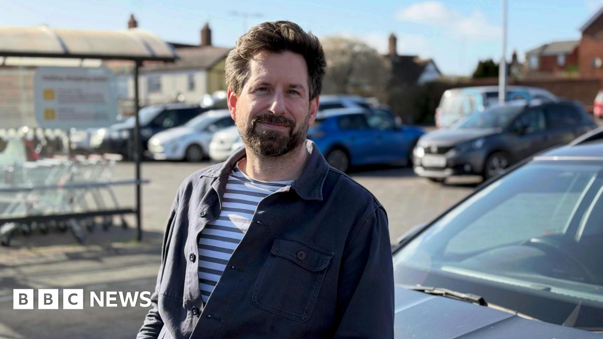 Gareth Wild stands in front of his car at the car park of his local supermarket. He looks at the camera and has dark brown hair and a beard. He is wearing a striped t-shirt underneath a navy blue shirt.