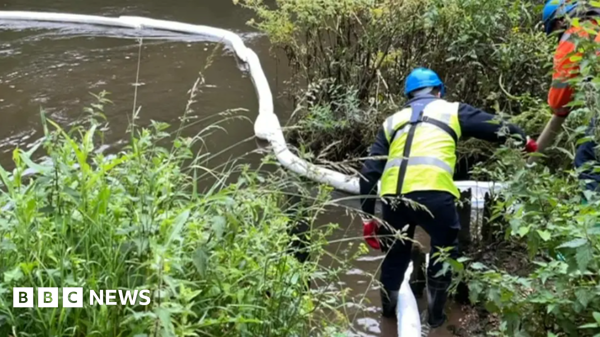 A helmeted worker stands in a river next to a floating bund which is being used to contain oil