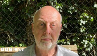 Steve Clare has short grey hair and is looking at the camera. He has a short white beard and he is sitting outside on a bench. There is a hedge behind him through a fence.