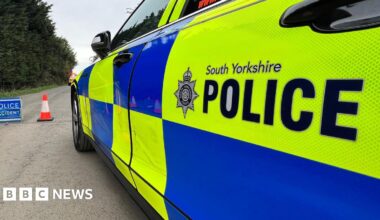 A South Yorkshire Police car with bright yellow‑and‑blue markings parked on a road, with a traffic cone and a blue “Police Accident” sign set up nearby, against a backdrop of trees.