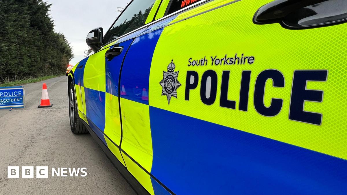 A South Yorkshire Police car with bright yellow‑and‑blue markings parked on a road, with a traffic cone and a blue “Police Accident” sign set up nearby, against a backdrop of trees.