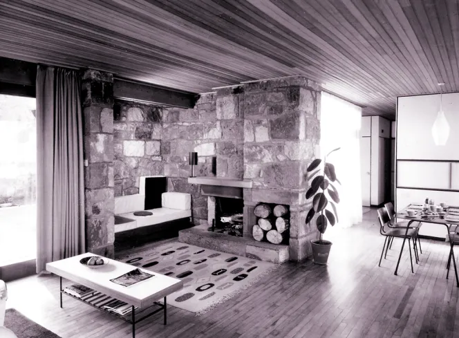 Black and white photo of the Tomlinson House's living room with a stone fireplace, wood floors, and a dining table in the background.