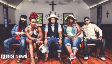A promotional photo of Arrested Development. Three men and two women sit on a bench inside a small chapel, which has crosses on the wall and stained glass windows. They all pose and look at the camera with serious expressions.  They wear denim jeans and two have sunhats on.
