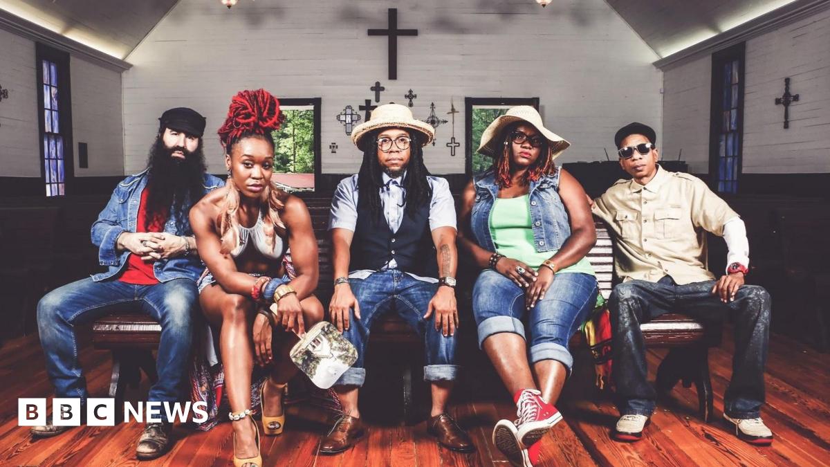 A promotional photo of Arrested Development. Three men and two women sit on a bench inside a small chapel, which has crosses on the wall and stained glass windows. They all pose and look at the camera with serious expressions.  They wear denim jeans and two have sunhats on.