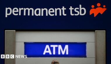 A close up of an atm. Lettering states permanent tsb ATM. A red logo shows three people. A reflection in a small circle shaped mirror displays a street.