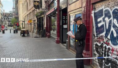 A police officer stands behind police tape on a city street in central London, reading notes outside a closed shop with graffiti on its shutter, while nearby streets are quiet and cordoned off.