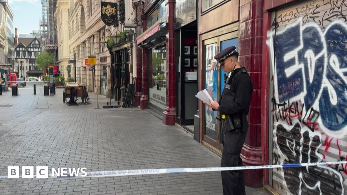 A police officer stands behind police tape on a city street in central London, reading notes outside a closed shop with graffiti on its shutter, while nearby streets are quiet and cordoned off.