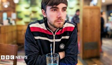 Nathan sitting in a pub with a drink. He is wearing a blue tracksuit top and has his arms crossed. He has a short beard and dark brown hair.