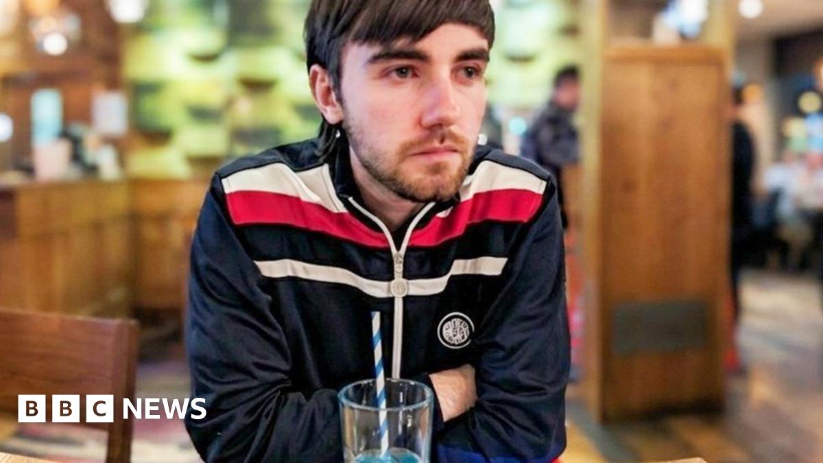 Nathan sitting in a pub with a drink. He is wearing a blue tracksuit top and has his arms crossed. He has a short beard and dark brown hair.