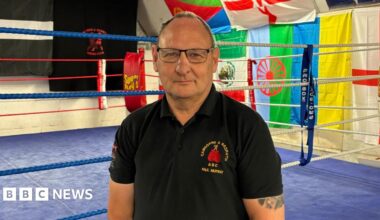 A man with a mostly bald head and wearing glasses in a dark polo shirt standing in front of a boxing ring.