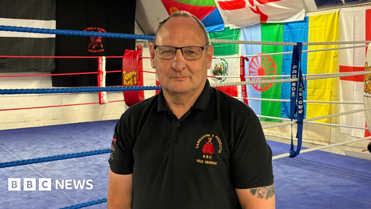 A man with a mostly bald head and wearing glasses in a dark polo shirt standing in front of a boxing ring.