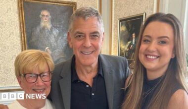 Three people, two women and George Clooney, posing together indoors on a patterned carpet, standing in front of framed artwork, dressed smartly with one person resting an arm around another.