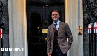 A smiling man stood outside the front door of Number 10 Downing Street. He is wearing a brown suit jacket with black waistcoat underneath, white shirt and brown tie. Attached to some black railings are St George's flags.